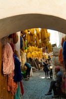 A narrow, cobblestone alleyway is adorned with bright, colorful skeins of yarn in shades of yellow, red, blue, and orange. The yarn hangs densely from overhead and against the walls. People are walking and inspecting the area, with some sitting and others standing under the shade of the archway.