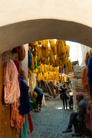 A narrow, cobblestone alleyway is adorned with bright, colorful skeins of yarn in shades of yellow, red, blue, and orange. The yarn hangs densely from overhead and against the walls. People are walking and inspecting the area, with some sitting and others standing under the shade of the archway.