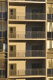 A series of balconies stacked vertically on a building facade, each featuring a small door and surrounded by metal railings. The facade appears to be in shadow, with sunlight casting across part of the image. The windows adjacent to the balconies are part of a glass-walled section of the building, reflecting muted light.