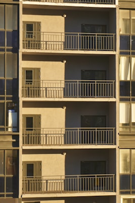 A series of balconies stacked vertically on a building facade, each featuring a small door and surrounded by metal railings. The facade appears to be in shadow, with sunlight casting across part of the image. The windows adjacent to the balconies are part of a glass-walled section of the building, reflecting muted light.