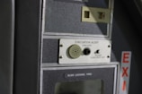 A close-up of a control panel featuring an evacuation alert section with a speaker, button, and label. The panel has a textured surface with the word 'EXIT' visible on the side in red letters.