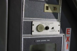A close-up of a control panel featuring an evacuation alert section with a speaker, button, and label. The panel has a textured surface with the word 'EXIT' visible on the side in red letters.
