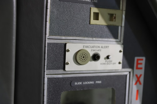 A close-up of a control panel featuring an evacuation alert section with a speaker, button, and label. The panel has a textured surface with the word 'EXIT' visible on the side in red letters.