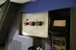 The image features a panel with a series of buttons and a handset. The panel includes labels such as 'CALL', 'FLIGHT DECK', and 'CANCEL' alongside three buttons of different colors and shapes. There is a plaque with numbers and text attached to the structure, and the lighting suggests it is located in a dimly lit environment.