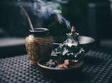 A ceramic incense holder with an intricate design releases cascading smoke. Next to it, a decorative cup with a metal straw, possibly for mate, features intricate motifs. Both are placed on a textured dark surface, creating a serene and contemplative atmosphere.