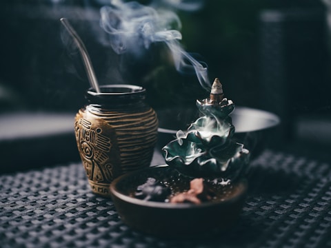 A ceramic incense holder with an intricate design releases cascading smoke. Next to it, a decorative cup with a metal straw, possibly for mate, features intricate motifs. Both are placed on a textured dark surface, creating a serene and contemplative atmosphere.