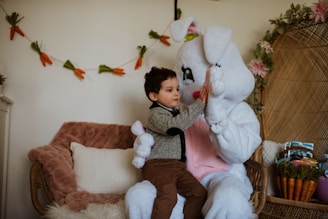 A young child sits next to a person dressed in a white bunny costume, giving them a high-five. The setting features cozy furniture with soft cushions and a rabbit-themed garland of carrots in the background. The area is decorated with flowers and has a warm, inviting atmosphere.