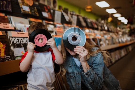 A child and an adult are in a record store, playfully holding vinyl records up to their faces. The background is filled with shelves of records and albums. The child is wearing a white and red shirt, while the adult is in a denim jacket.
