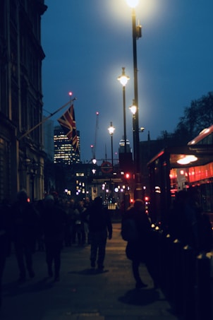 An evening city street scene featuring illuminated street lamps, a British flag, and a busy sidewalk with silhouettes of people. In the background, there are skyscrapers and a London Underground sign for Westminster Station.