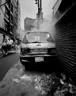 A damaged car emitting smoke is parked closely against a brick wall in a narrow urban alley. Nearby, a rickshaw with two men is passing through the street. The overall atmosphere is gritty, with visible litter on the ground and wires hanging overhead.