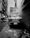A damaged car emitting smoke is parked closely against a brick wall in a narrow urban alley. Nearby, a rickshaw with two men is passing through the street. The overall atmosphere is gritty, with visible litter on the ground and wires hanging overhead.
