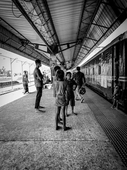 A group of people stand on a train platform, interacting with each other. The platform is covered with a high, structured roof, and a train is visible on the right side. Children and adults appear to be waiting or socializing.