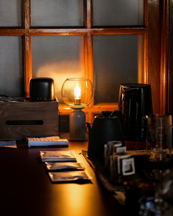A warmly lit scene with a glowing lamp sitting on a table next to a wooden box. There are various objects including a calculator, electric kettle, and stacked packets. The light casts a soft amber glow, with reflections on the table surface.