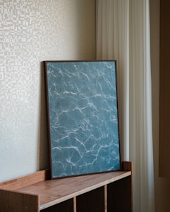 A framed piece of art with a marble-like pattern is resting on a wooden shelf. The art features a swirling blue design with white veining. It leans against a textured beige wall and is partially framed by white drapes.