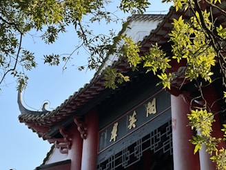 A traditional architectural structure features ornate roof tiles and decorative elements, partially obscured by lush green tree branches and leaves. The sunlight casts highlights on the building, creating a harmonious blend with the natural elements.
