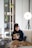 A woman is sitting comfortably on a sofa in a modern living room, looking at her phone. She is wearing a dark sweater with a graphic design and a brown blanket draped over her legs. A stylish lamp stands beside her, and a shelf with decorative plants is visible in the background.