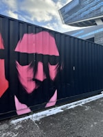 A pink stylized portrait of a face is painted on the side of a black shipping container, located outdoors. The background features a modern architectural building with glass facades under a partly cloudy blue sky. The ground is covered in a light dusting of snow.