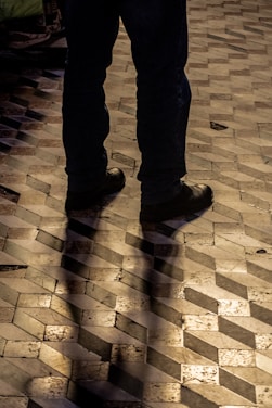 The image captures the lower half of a person standing on a geometric patterned floor. The flooring consists of rectangular tiles in shades of brown and beige, creating a 3D cube illusion. The person is wearing dark pants and black shoes, and their shadow is cast on the floor due to the lighting.