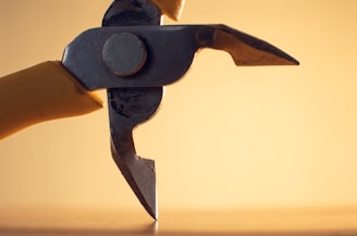A close-up view of a metallic tool, possibly wire cutters, set against a warm, soft yellow background. The tool has sharp, distinct blades and a yellow handle. The lighting creates a dramatic shadow and highlights the texture of the metal.
