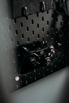 A black gaming controller is mounted on a perforated metal board, with hooks and a headset also visible. The environment appears organized and functional, with a modern aesthetic.