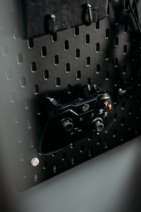 A black gaming controller is mounted on a perforated metal board, with hooks and a headset also visible. The environment appears organized and functional, with a modern aesthetic.