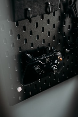 A black gaming controller is mounted on a perforated metal board, with hooks and a headset also visible. The environment appears organized and functional, with a modern aesthetic.