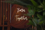 Soft lighting illuminates a draped fabric backdrop with the words 'Better Together' elegantly written in script. Green foliage partially obscures the view, adding a natural element to the composition.