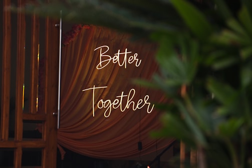 Soft lighting illuminates a draped fabric backdrop with the words 'Better Together' elegantly written in script. Green foliage partially obscures the view, adding a natural element to the composition.