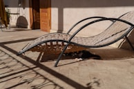 A woven lounger is positioned in an outdoor space with sunlight casting strong shadows across the floor and the chair. A black and white cat is lying underneath the lounger, partially visible and enjoying the shade. The surroundings include a doorway with a wooden door and a small table with a plant.