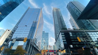 Tall, modern skyscrapers with glass facades stretch into the blue sky. The buildings reflect the surrounding cityscape, creating a dynamic urban scene. Street signs and a traffic light are visible, and a bank logo is prominently displayed, indicating a financial district.