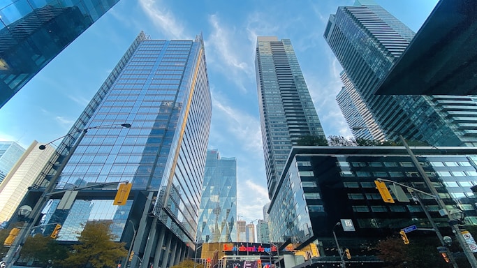 Tall, modern skyscrapers with glass facades stretch into the blue sky. The buildings reflect the surrounding cityscape, creating a dynamic urban scene. Street signs and a traffic light are visible, and a bank logo is prominently displayed, indicating a financial district.