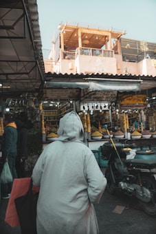 A bustling market scene with a person dressed in a long robe walking towards various stalls. The stalls are filled with large piles of brightly colored spices. Above the market, a building with a sign reading 'Caf&eacute;' has an open terrace. A scooter is parked on the side near the market.