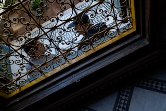 A wrought iron grill with intricate patterns is seen from an angle. Beyond the grill, there is a tiled courtyard with geometric patterns and plants visible. Two people are partially visible through the grill, possibly engaged in conversation.