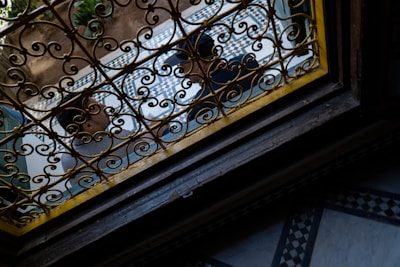 A wrought iron grill with intricate patterns is seen from an angle. Beyond the grill, there is a tiled courtyard with geometric patterns and plants visible. Two people are partially visible through the grill, possibly engaged in conversation.