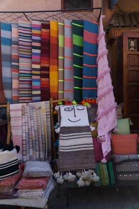 A display of colorful woven textiles in various patterns and colors is arranged against a backdrop. The textiles include stripes and solid colors, suggesting they are artisanal blankets or rugs. One piece features a smiling face design, and there are several folded textiles stacked below.