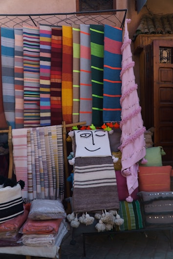 A display of colorful woven textiles in various patterns and colors is arranged against a backdrop. The textiles include stripes and solid colors, suggesting they are artisanal blankets or rugs. One piece features a smiling face design, and there are several folded textiles stacked below.