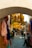 A narrow cobblestone alley is adorned with vibrant skeins of yarn hanging overhead and on the walls. People are walking and interacting in the marketplace, with one person sitting in the foreground, creating a lively atmosphere.