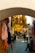 A narrow cobblestone alley is adorned with vibrant skeins of yarn hanging overhead and on the walls. People are walking and interacting in the marketplace, with one person sitting in the foreground, creating a lively atmosphere.