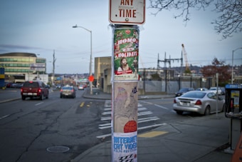 An urban street view featuring a utility pole adorned with colorful posters. The posters advertise events, including one with festive decorations and a character dressed as Santa. Nearby, there are several vehicles on the road, buildings in the background, and a telephone booth on the sidewalk.