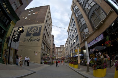 A lively urban street scene with tall buildings lining both sides. There are string lights hanging overhead, and colorful planters with flowers on the sidewalk. People are gathered in small groups, some sitting and enjoying meals at outdoor restaurants.