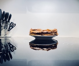 A stack of crispy, golden-brown flatbreads is placed on a black plate. The setting is modern with an array of black-handled kitchen knives and scissors organized in a clear block on the left. The reflective surface beneath the plate adds an elegant touch, creating a subtle mirror effect.