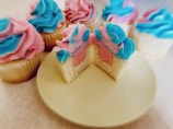 Several cupcakes are arranged with brightly colored frosting in pastel shades of blue and pink. One cupcake has been cut in half to reveal a two-toned interior matching the frosting colors. The cupcakes are displayed on a light-colored surface.