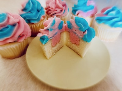 Several cupcakes are arranged with brightly colored frosting in pastel shades of blue and pink. One cupcake has been cut in half to reveal a two-toned interior matching the frosting colors. The cupcakes are displayed on a light-colored surface.