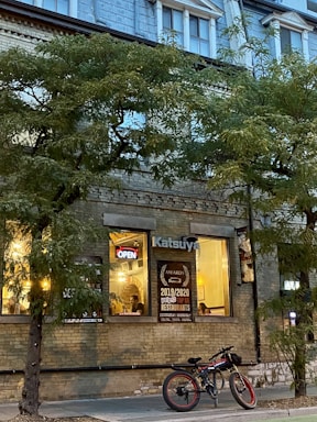 A cozy restaurant setting with warm lights shining through large windows displaying awards and an 'Open' sign. Trees line the sidewalk, and a bicycle is parked in front. The building features a bricked facade with upper apartment windows.
