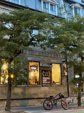 A cozy restaurant setting with warm lights shining through large windows displaying awards and an 'Open' sign. Trees line the sidewalk, and a bicycle is parked in front. The building features a bricked facade with upper apartment windows.