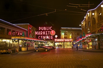 A vibrant and bustling market area at night, adorned with festive lights and decorations. The architecture is reminiscent of a historic district, featuring a prominent neon sign that reads 'PUBLIC MARKET CENTER'. Several Christmas trees and holiday lights add to the festive atmosphere. The street is paved with cobblestones, and a few cars are parked along the curb. People are seen walking towards a bright 'FARMERS MARKET' sign in the background.