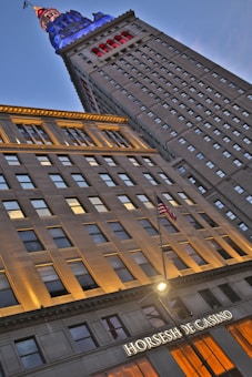 Tall building with illuminated top in blue and red lights, seen from a low angle. The sign 'Horseshoe Casino' is visible near the entrance. An American flag is also visible in the foreground. The building has numerous windows and a street light is present in front of the casino.