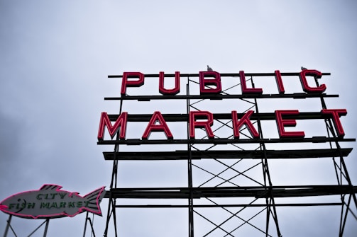 A large sign with the words 'PUBLIC MARKET' in bold red neon letters, supported by a metal framework. To the left, there is an additional sign shaped like a fish with the text 'CITY FISH MARKET'. The background is a cloudy sky, adding a moody atmosphere to the scene.