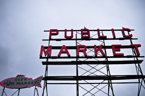 A large sign with the words 'PUBLIC MARKET' in bold red neon letters, supported by a metal framework. To the left, there is an additional sign shaped like a fish with the text 'CITY FISH MARKET'. The background is a cloudy sky, adding a moody atmosphere to the scene.