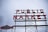 A large sign with the words 'PUBLIC MARKET' in bold red neon letters, supported by a metal framework. To the left, there is an additional sign shaped like a fish with the text 'CITY FISH MARKET'. The background is a cloudy sky, adding a moody atmosphere to the scene.